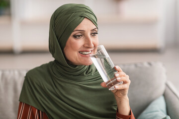 Wall Mural - Cheerful Senior Muslim Lady In Hijab Drinking Water At Home