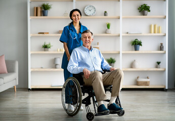 Full length portrait of older disabled man in wheelchair and his young caregiver smiling at camera in retirement home