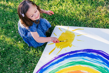 Little girl 2-4 years old paints rainbow and sun on large sheet of paper, sitting on green lawn