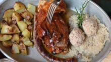An Appetizing Pan With Some Trentino Specialties: Baked Pork Shank, Roasted Potatoes, Sausage Dumplings And Sauerkraut. Concept Of Typical And Tasty Italian Food. A Fork Touches The Shin.