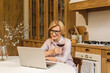 © denis_vermenko - Portrait of smiling mature senior woman holding glass of wine while using laptop on kitchen table. Freelance working at home concept.
