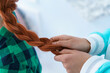 A friend is braiding a braid for a red-haired girl. Hairstyle for a cute young woman