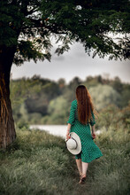 A Girl With Long Hair In A Green Dress With A Hat Goes To The River, View From The Back. Summer, Park, Walks In Nature And Fresh Air, Photo Session
