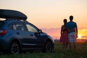 Happy couple relaxing beside their SUV car during honeymoon road trip at sunset. Young man and woman enjoying time together travelling by vehicle.