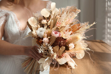 Bride holding beautiful dried flower bouquet near window at home, closeup