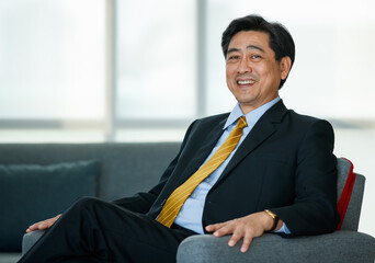 50s Asian senior executive businessman entrepreneur wearing formal suit with luxury necktie, smart sitting on sofa in indoor office, smiling with confidence, success and gladness with copy space.