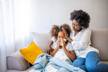 Wall Mural - Mother helping daughter blowing her nose on the sofa at home. Ill African American girl blowing a nose at home while her mother is consoling her. 