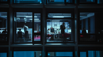 Poster - Shot from Outside the Window: Businessmen and Businesswomen Working in the Office. Managers and Specialists Doing Financial Business in the Evening. Employees Work on Computers and Delegate Tasks.