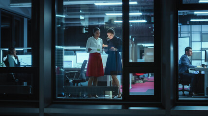 Poster - Shot from Outside: Two Businesswomen Working on a Tablet Computer in Office. Managers Standing by the Window. Employees Talk about Financial and Business Development. Colleagues Work Behind Them.