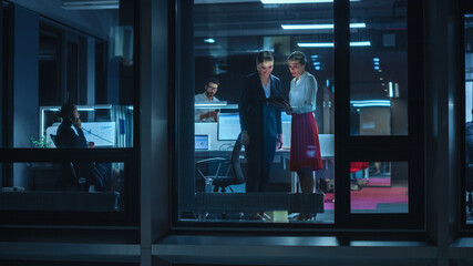 Poster - Shot from Outside: Two Businesswomen Working on a Tablet Computer in Office. Managers Standing by the Window. Employees Talk about Financial and Business Development. Colleagues Work Behind Them.