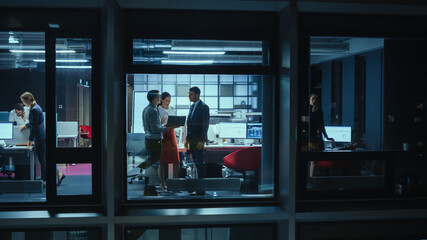 Shot from Outside the Window: Daily Meeting in the Office. Female Manager Explaining New Business Strategy to Young Financial Specialists on a Laptop Computer while Standing Next to a Window.