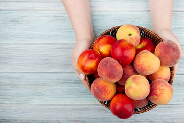 Poster - top view of female hands holding a wicker basket with fresh ripe nectarines and peaches at rustic wooden background with copy space