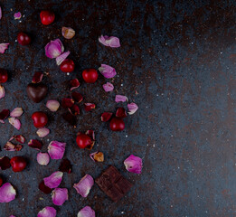 Poster - top view of rose petals with heart shaped chocolate candies and red cherries on black background with copy space