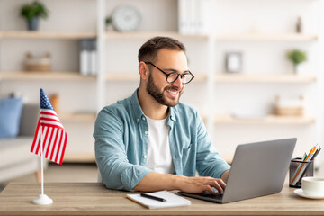 Poster - Happy young guy with flag of the US working at desk with laptop in home office