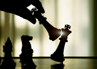 Wall Mural - Hand holding black king attack white king of chess silhouettes battle on board. Black king are leader to fight with teamwork to victory. Leader,strategy and checkmate concept for success.