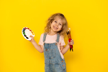 Blonde curly girl on a yellow background smiles, with paints and tassels in her hands, dressed in a denim jumpsuit and pink T-shirt