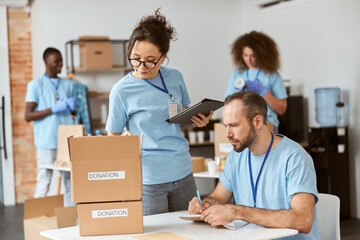 Wall Mural - Man and woman working together on donation project indoors. Team of volunteers in blue uniform making notes, calculating, sorting and packing items in cardboard boxes