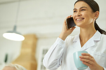 Wall Mural - Smiling female doctor using smartphone and drinking hot drink