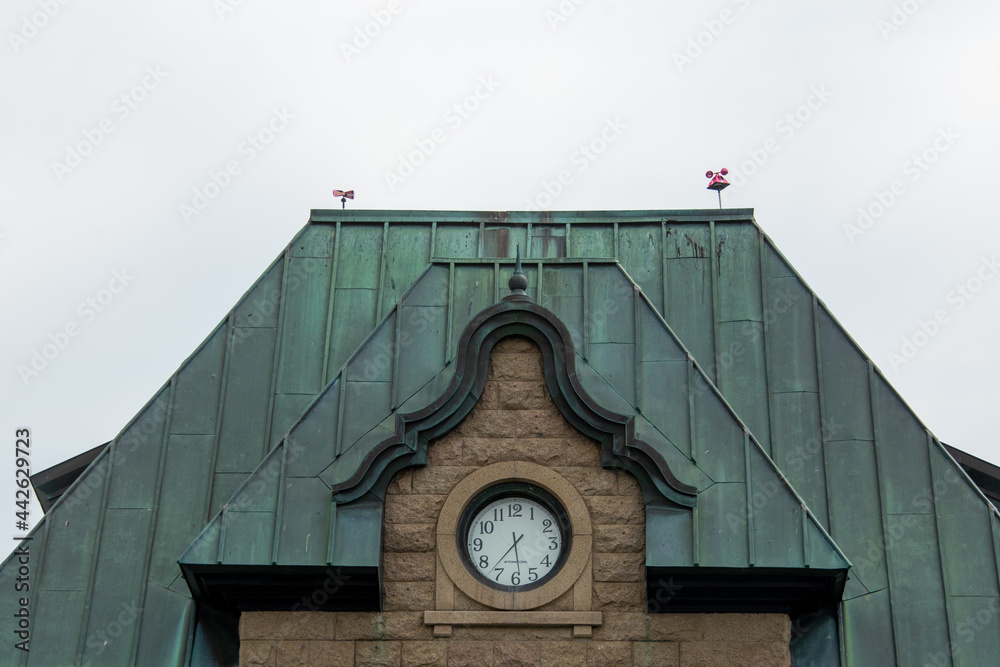The exterior of a vintage brown brick building with an oxidized copper roof.  The front of the building has a white round clock with black numbers. The grey sky is in the background.