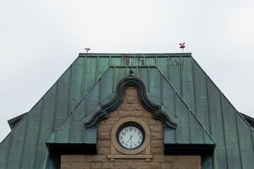 The exterior of a vintage brown brick building with an oxidized copper roof.  The front of the building has a white round clock with black numbers. The grey sky is in the background.