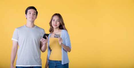 Wall Mural - Asian couple holding credit card and smartphone while looking at a camera isolated on yellow background