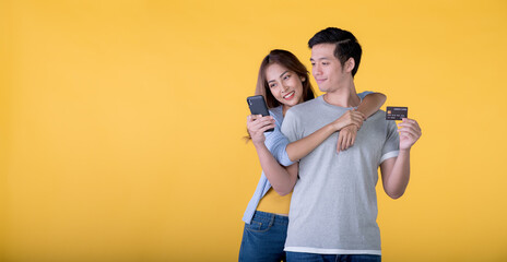 Wall Mural - Excited Asian couple holding credit card and smartphone feeling excited while looking at a mobile phone isolated on colour background