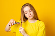 Sad and unhappy teen girl cutting her hair with scissors while standing on yellow background. Young student experiments with her hairstyle at home.