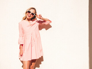 Young beautiful smiling hipster woman in trendy summer pink dress. Sexy carefree woman posing near white wall in the street at sunset. Positive model outdoors.Happy and cheerful.Shows peace sign