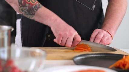 Wall Mural - A male chef is a professional in an apron preparing a salad of vegetables in the kitchen of the restaurant. The cook cuts the raw carrot with a knife on a cutting board. Close-up.