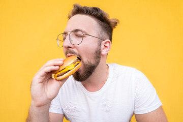 Creative closeup portrait of a young handsome bearded guy with glasses in a white t-shirt tastes a hamburger from a fast food on the yellow background.