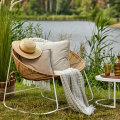 Stylish composition of outdoor garden on the lake with design rattan armchair, coffee table, plaid, pillows, drinks and elegant accessories. Summer chillout mood.