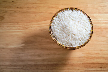 overhead flat lay shot of raw Thai's jasmine rice in Thai's traditional style bamboo's bowl on wooden dinner table with light and shade and some copy space