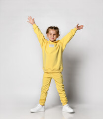 Wall Mural - Full length portrait of a little girl dressed in a bright yellow sports suit. Cute child posing on a white background with arms raised. Concept of active childhood.