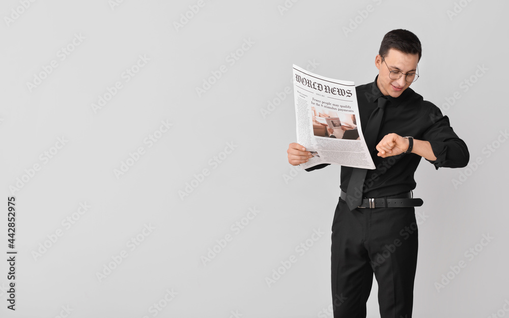 Young businessman with newspaper looking at wristwatch on light background
