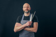 Bald male butcher or fishmonger in black t shirt and classic black and white apron on dark background. Meat industry. Holding knife and metal steel. Man in his 40s, grey beard, smile on his face