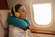© New Africa - Young woman with travel pillow resting while listening to music in airplane during flight