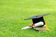 A mortarboard and graduation scroll, tied with red ribbon, on a stack of books on green grass springtime in the outdoor park. Concept education congratulation.
