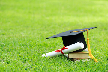 A mortarboard and graduation scroll, tied with red ribbon, on a stack of books on green grass springtime in the outdoor park. Concept education congratulation.