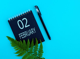 February 2th. Day 2 of month, Calendar date. Black notepad sheet, pen, fern twig, on a blue background. Winter month, day of the year concept