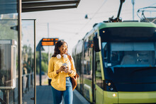 Attractive African American Female Traveler Or Student Waiting For Public Transport Standing On Bus Stop Outdoor At Sunny Morning With Transport On Blurred Background.