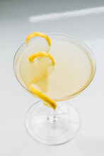 Yellow Cocktail In Coupe Glass With Long Curly Lemon Twist Against White Background