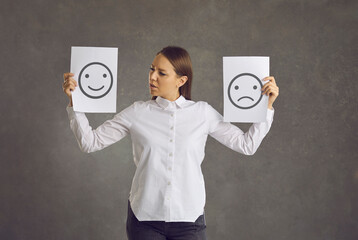 Confused young woman holding two sheets of paper with happy and unhappy emoticons