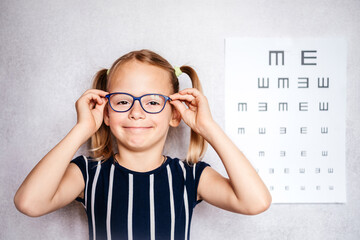 Wall Mural - Happy little girl wearing eyeglasses taking eyesight test before school with blurry eye chart at the background, kindergarten and school medical check up