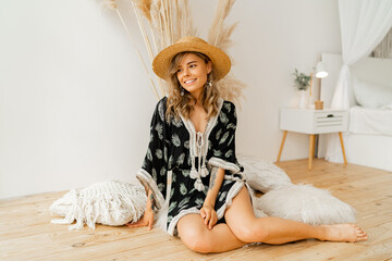 Wall Mural - Stylish  blond woman in straw hat and boho dress  posing in studio over white background  with pampas grass decor.  Pillows with macrame ornament. Summer trends.