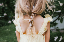 Portrait Of A Little Blonde Girl In A Yellow Dress With Braid With Braided Lilac Flowers Standing Near A Blooming Lilac In Spring, The Concept Of A Happy Childhood And School Holidays