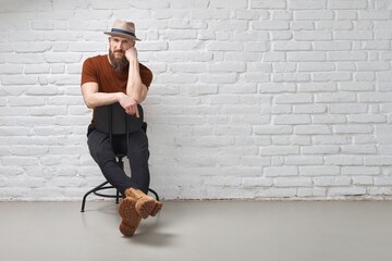 Wall Mural - Serious bearded man in hat sitting in front of white brick wall
arms crossed. Full length photo with copy space.