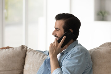 Wall Mural - Close up profile view of smiling man talking on phone sitting on cozy couch, happy positive young male speaking enjoying pleasant conversation with friend or girlfriend, making call by telephone