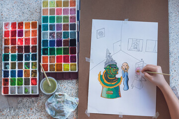 Top view of young girl's hand is painting art picture on paper with 2 watercolor trays and artistic things on white marble table