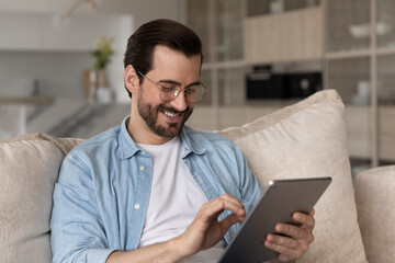 close up smiling man in glasses using tablet at home, sitting on cozy couch, happy young male lookin