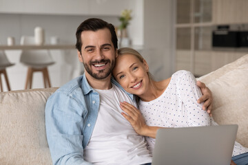 Wall Mural - Head shot portrait of happy romantic couple hugging, using laptop together, smiling wife and husband looking at camera, sitting relaxing on couch at home, enjoying leisure time with device